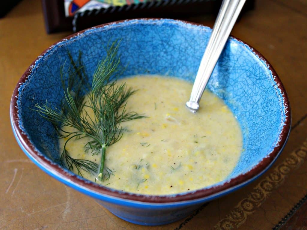 Summer Squash Chowder soup in a blue bowl with a sprig of dill and a spoon in the soup on a table.