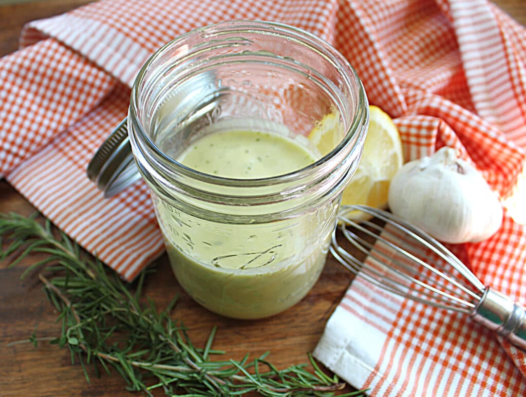 Close up of Dijon Italian Salad Dressing in a mason jar with garlic, lemon, whisk, rosemary