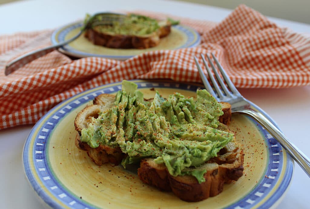 2 avocado toasts on yellow and blue plates with forks and an orange and white cloth napkin.