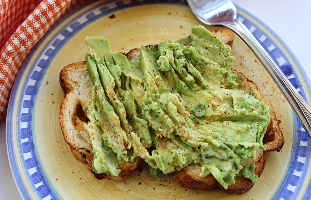 close up of prepared avocado toast on a yellow and blue plate with a fork.