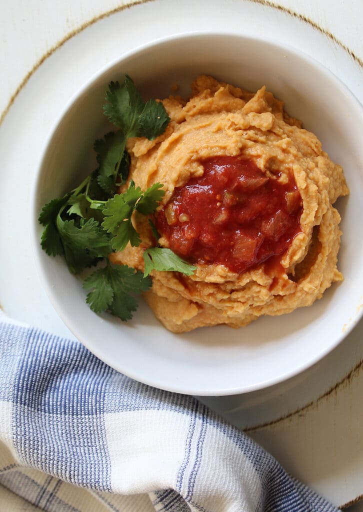 homemade hummus with hot salsa on top and cilantro leaves on the side. In a white bowl with and a blue and white towel.