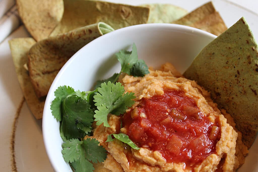 Homade hummus in a white bowl with cilantro and hot salsa on top. Served with homemade low carb spinach tortilla chips.