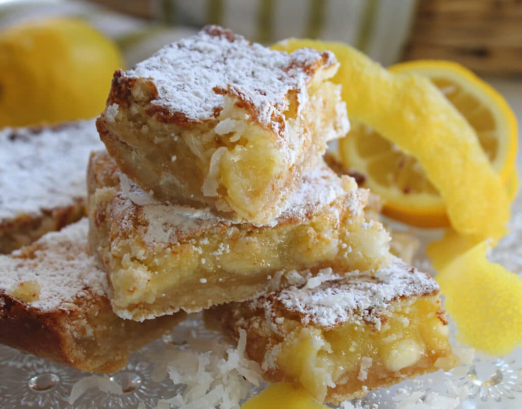 Up close stacked lemon bars with lemon shavings, lemons, coconut on a crystal serving tray.