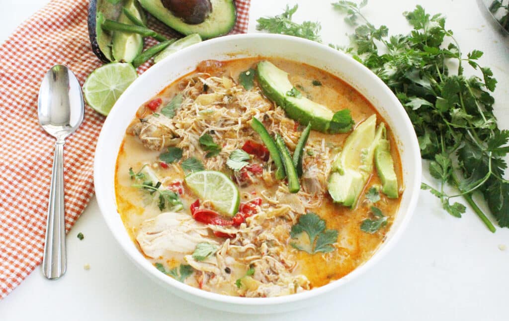 mexican chicken soup in a white bowl with avocado, cilantro and lime wedges