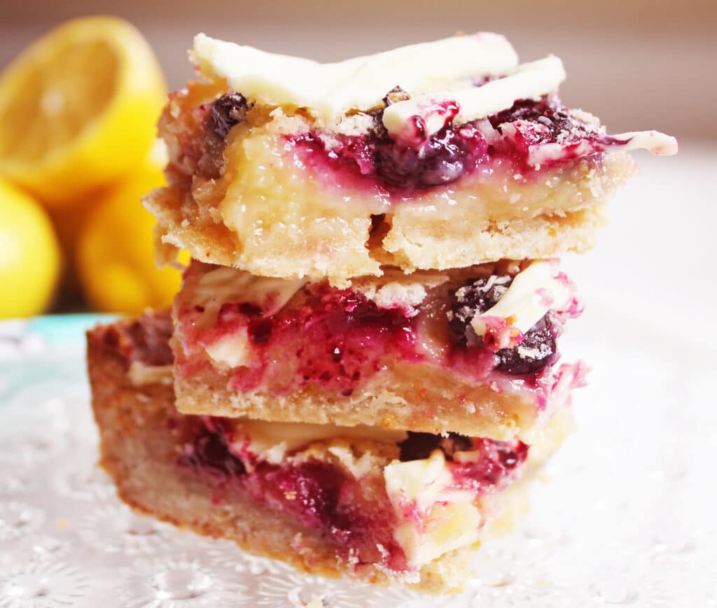 White Chocolate Blueberry Lemon Bars Stacked on a crystal serving plater with lemons in the background