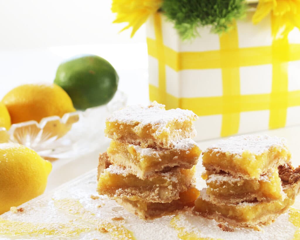 Stacked lemon bars on a crystal serving platter with lemons, limes and a flower vase.