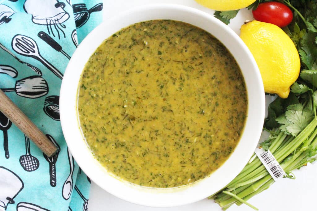 Overhead close up of Cilantro Dill Salad Dressing and Marinade in a white bowl with a blue towel and lemons, tomatoe and cilantro.