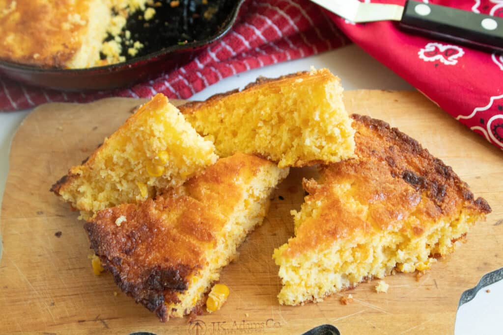 Cast iron skillet cornbread wedges losely stacked on a cutting board with a red cast iron skillet in the background