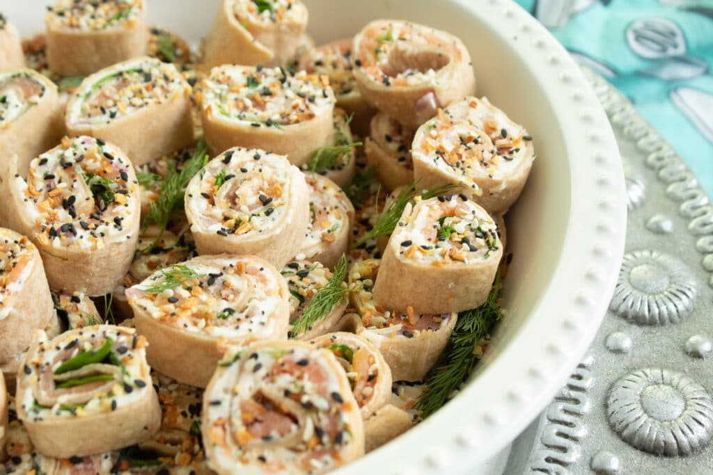 Many salmon everything pinwheels in a white shallow bowl on a silver dialed platter on a blue towel.