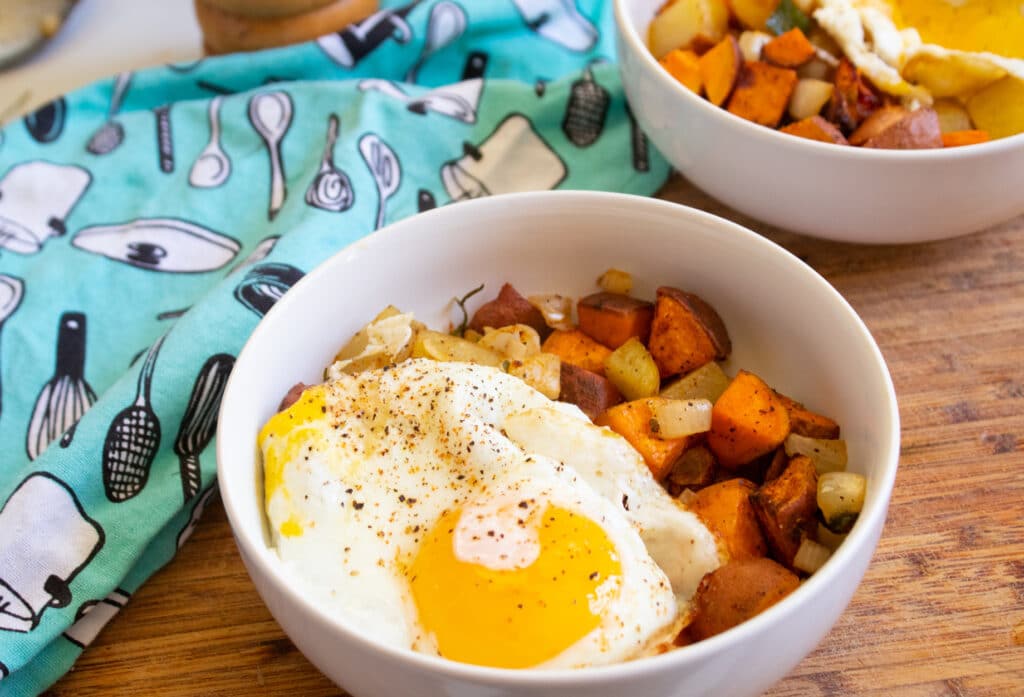 Two bowls of sweet potato hash with an egg on top on a cutting board with a teal towel.