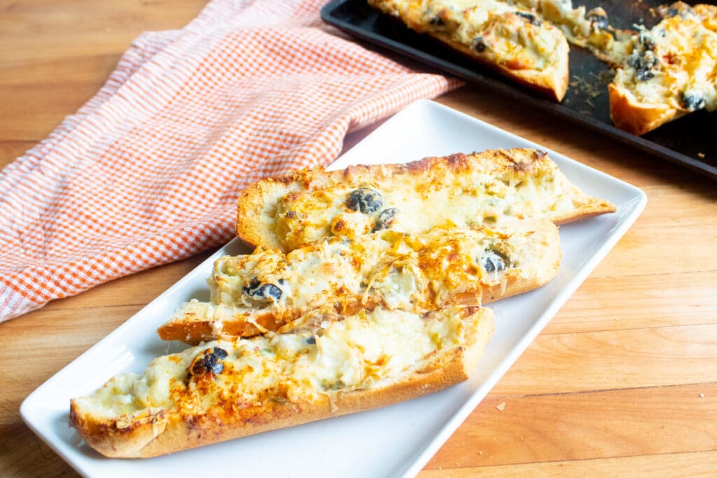 3 whole slices of artichoke cheese bread with olives on a white rectangle plate and a extra bread on a cookie tray