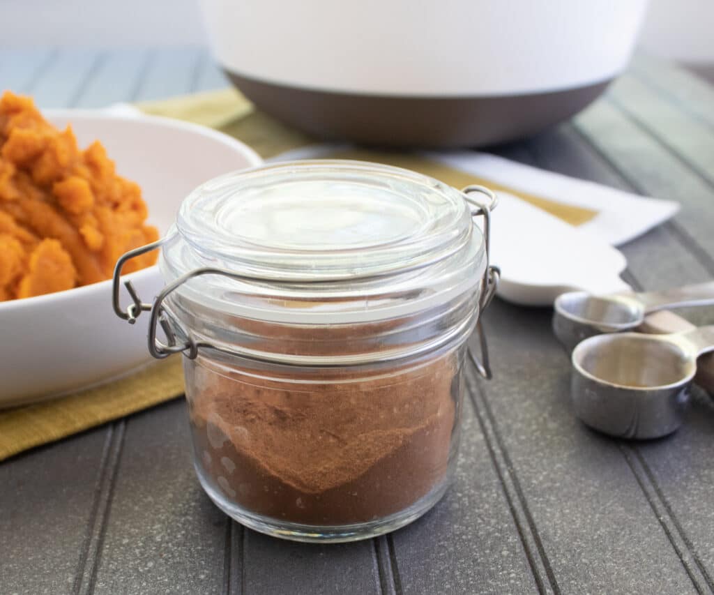 pumpkin pie spice in a glass jar with measuring spoons and pumpkin in the background