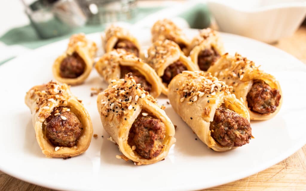 close up side view of cooked mini sausage rolls on a white plate.