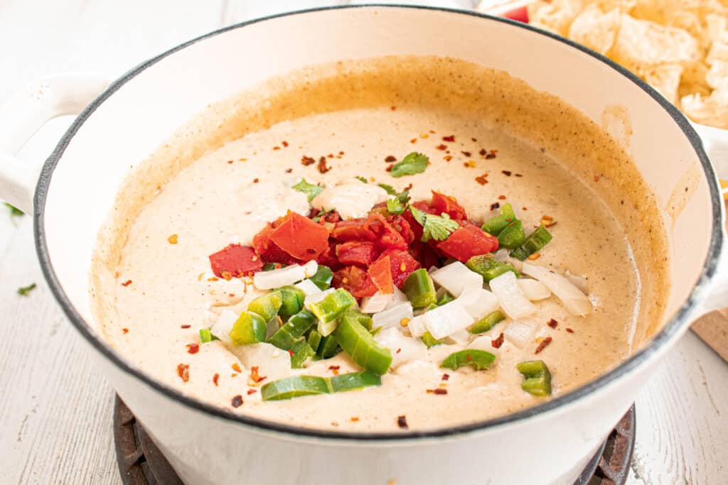 Finished white cauliflower Queso in a white enameled cast iron stock pot with peppers, onions, Rotel tomatoes, and red pepper on top.