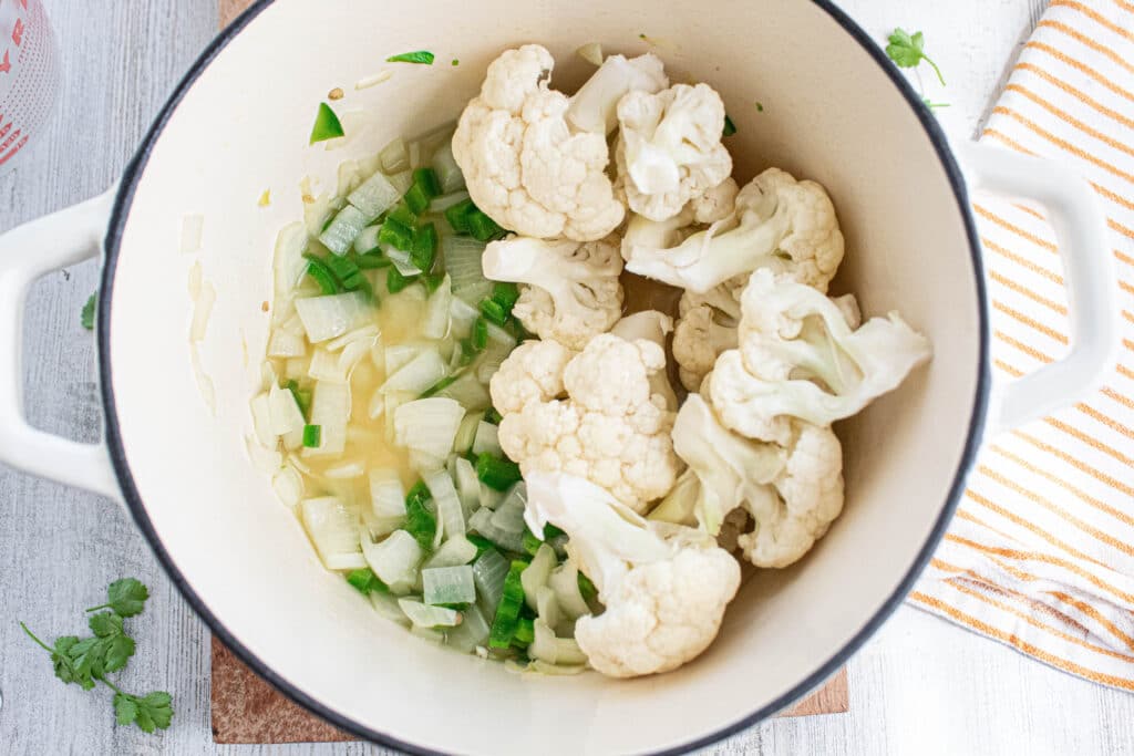 Sauteed peppers and onion in broth with raw cauliflower florets in a white dutch oven.