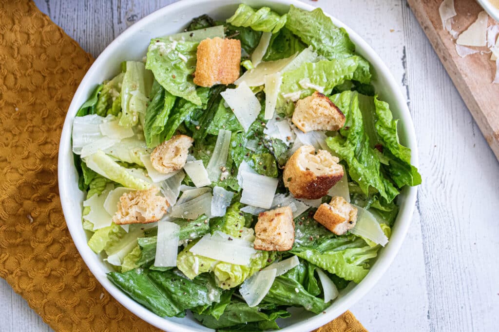 Caesar Salad with homemade croutons and shaved parmesan cheese in a white bowl on a mustard yellow towel.