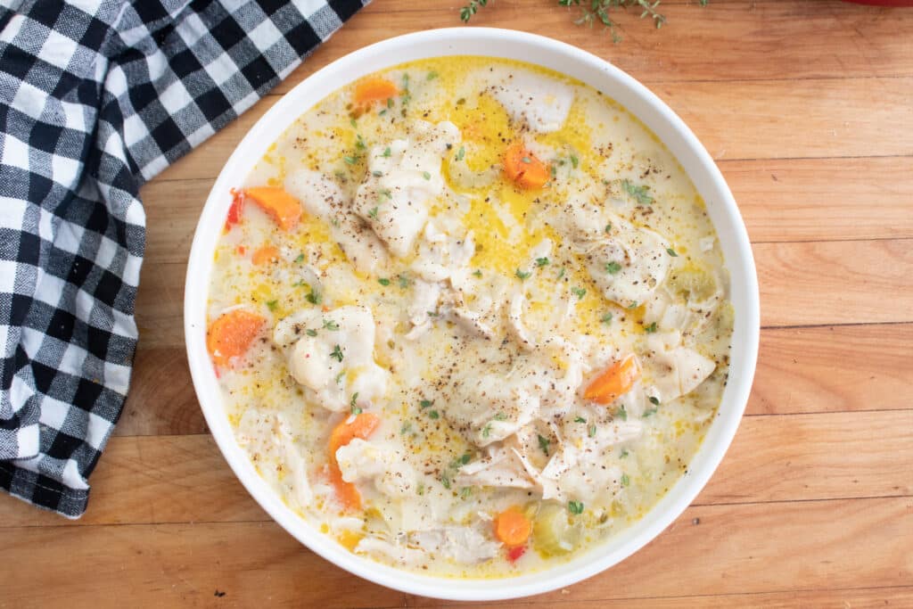 overhead of chicken and dumplings in a white bowl on a cutting board with a black and white checkered towel