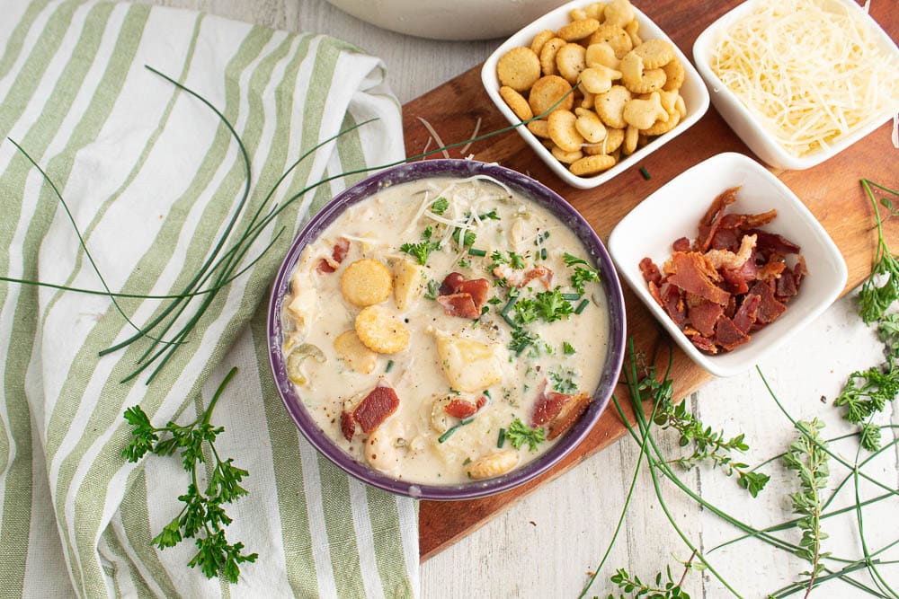 creamy and light new england clam chowder in a purple bowl in a brown board with toppings on a green and white striped tea towel