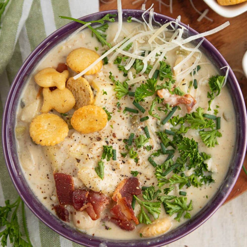 full purple bowl of creamy and light clam chowder with seasoned oyster crackers, bacon, parsley, chives on top