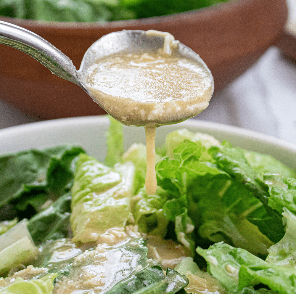 Serving ladle full of Caesar Dressing dripping onto a white bowl full of torn romaine lettuce.