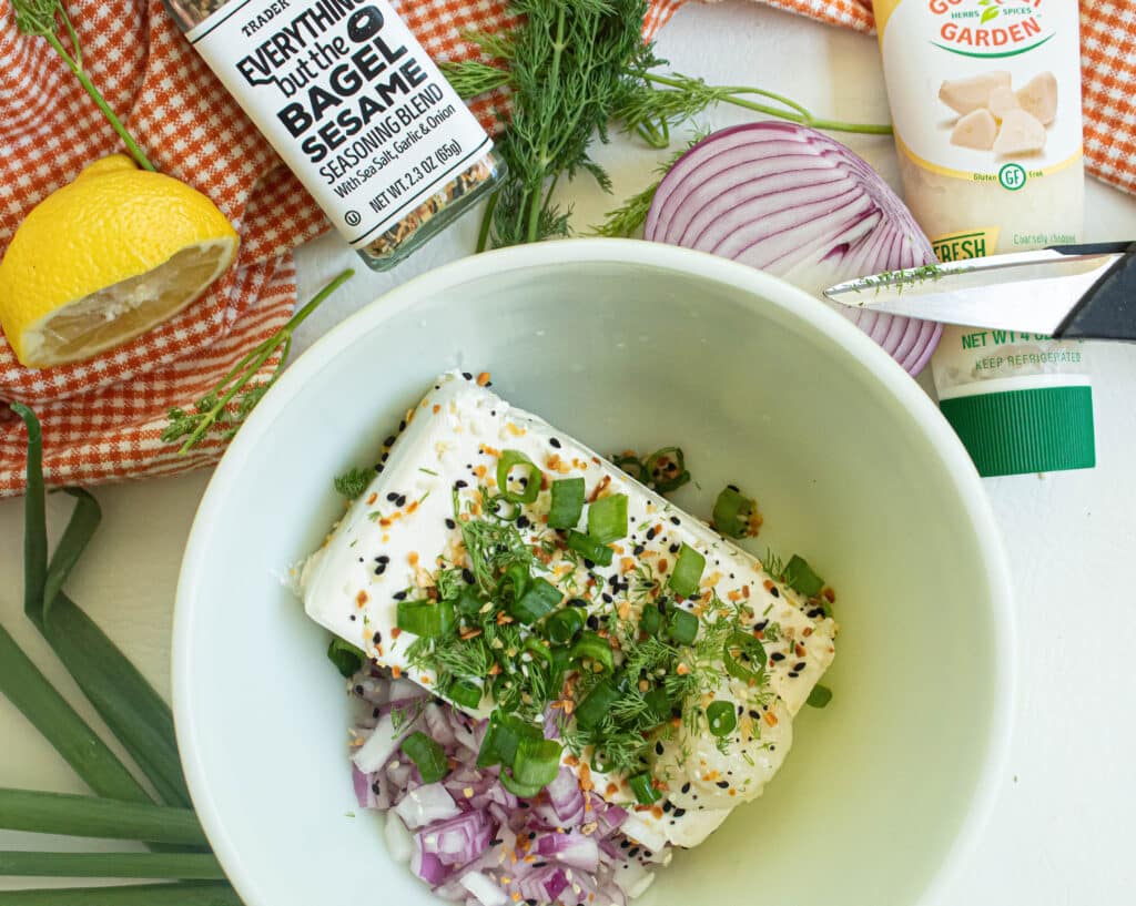 Cream cheese, red onions green onions, fresh dill, and Everything but the Bagel seasoning in a bowl. Red onion, garlic paste, seasoning and cut lemon on an orange and white checkered towel.