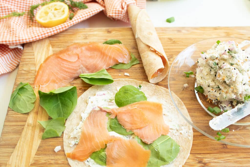 Tortilla with cream cheese mixture smeared on with a layer of fresh spinach and smoked salmon slices on top. Cream cheese mixture on a clear plate and a salmon slice and finished roll up on a wood cutting board.