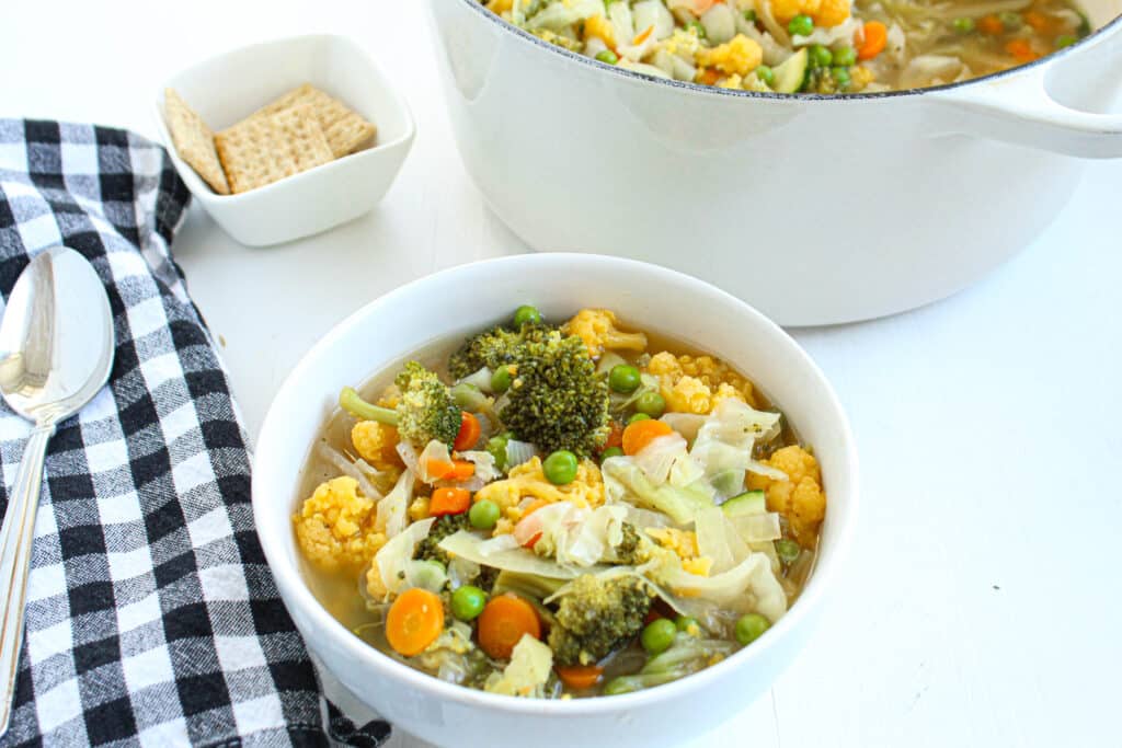 Easy vegetable soup in a white bowl with a black and white towel and spoon on the side. Large white dutch oven in the background full of easy vegetable soup.