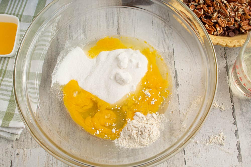 Eggs, sugar and flour in a large clear mixing bowl with pie shell and melted butter in the background.