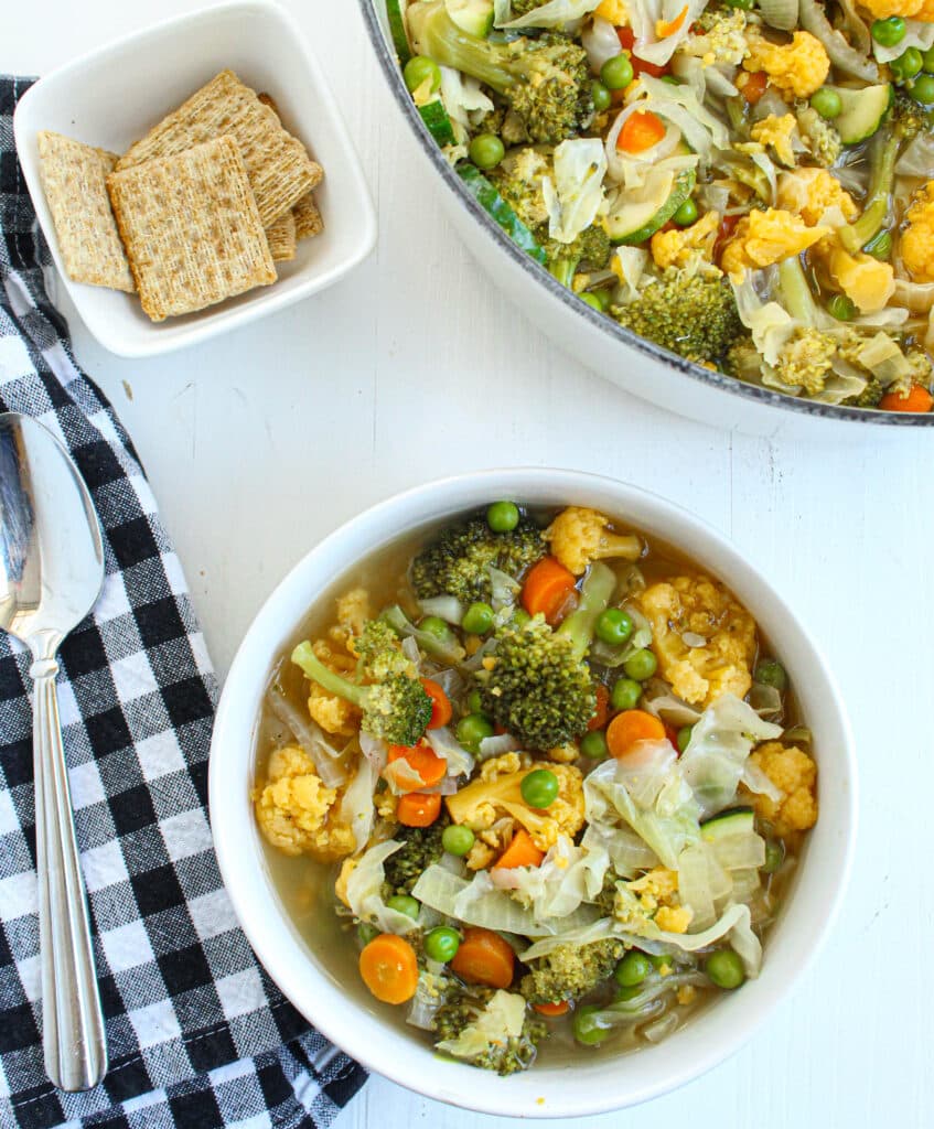 White bowl full of vegetable cabbage soup with a black and white towel and a spoon. Crackers in a white square bowl and a white dutch oven full of easy vegetable soup.