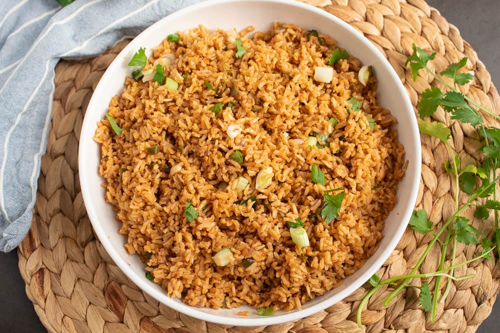 Mexican baked brown rice in a white bowl on a bamboo mate with a blue towel and cilantro and onion on top.