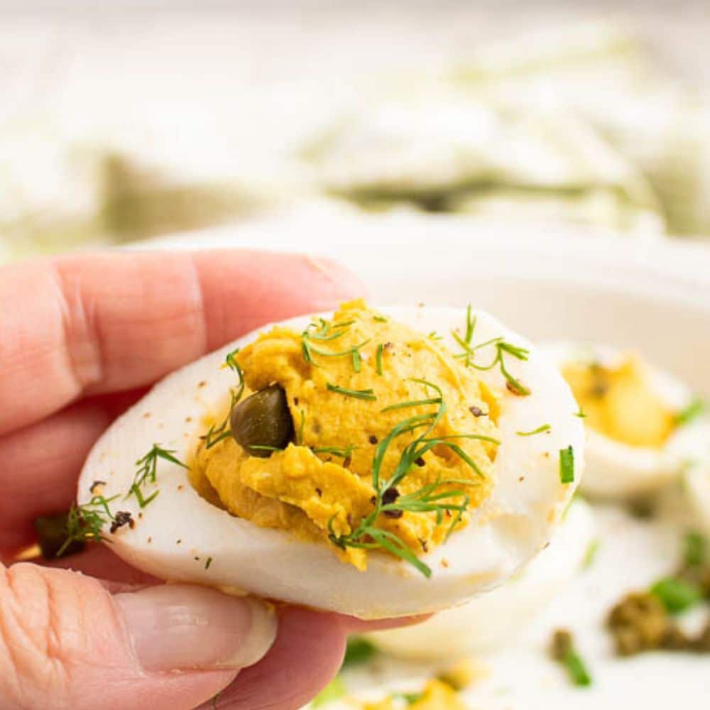 Single deviled egg with curry and dill toppings held by a hand over the plate of deviled eggs.