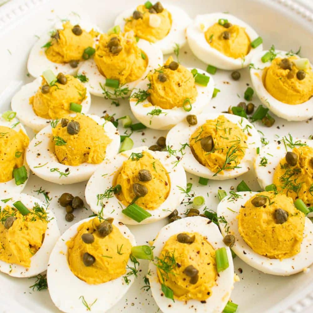 Plated deviled eggs with curry, dill, capers, and green onion on top in a white serving plate.