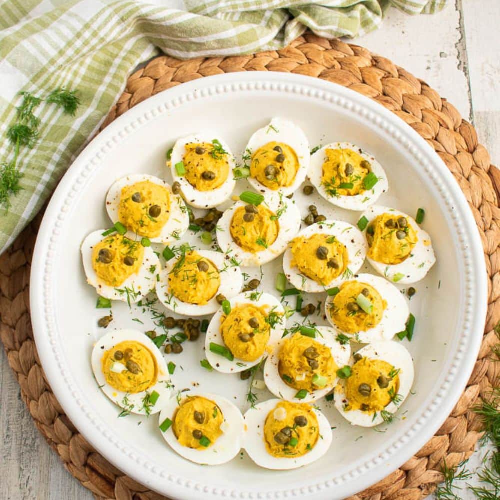 White plate filled with deviled eggs with curry and dill on a bamboo placemat on a white table with a green and white plaid towel.