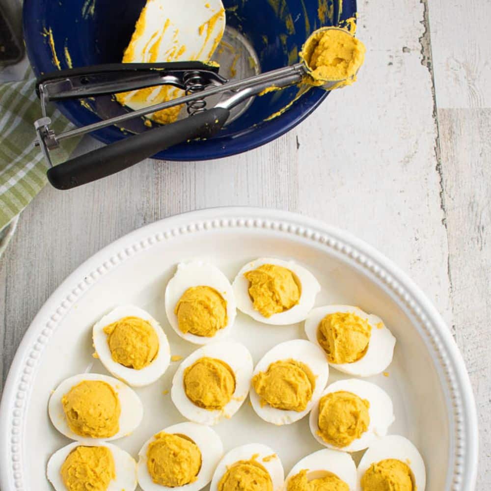 Hard boiled egg halves with yolk sauce mixture scooped inside halves in a white serving plate. Blue bowl with leftover mixture and small scoop utencil.