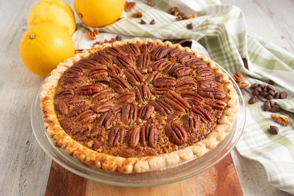 Whole Chocolate Orange Pecan Pie  in a glass pie plate on a wooden serving board with oranges, chocolate chips and pecans on a green and white plaid towel.