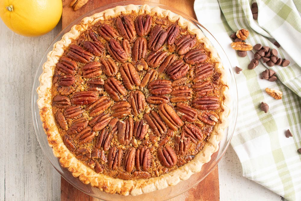 Chocolate Orange Pecan Pie with decorative pecans on top. On a grey table with chocolate chips and pecan pieces on a green and white plaid towel.