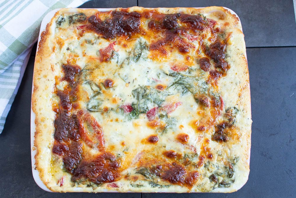 Cooked easy creamy white chicken lasagna with spinach and red peppers on a dark grey tile countertop and a green and white checked towel.