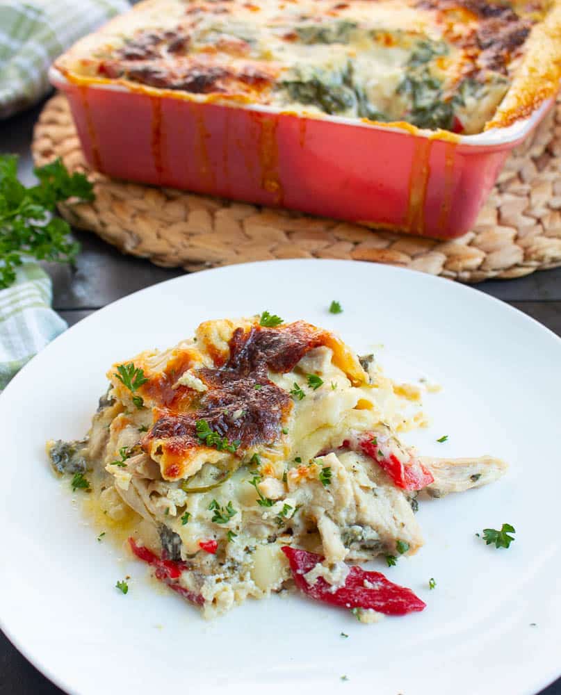 A single piece of Creamy White Chicken Lasagna with spinach and roasted red peppers on a white plate. The full dish is in the background on a bamboo placemat.