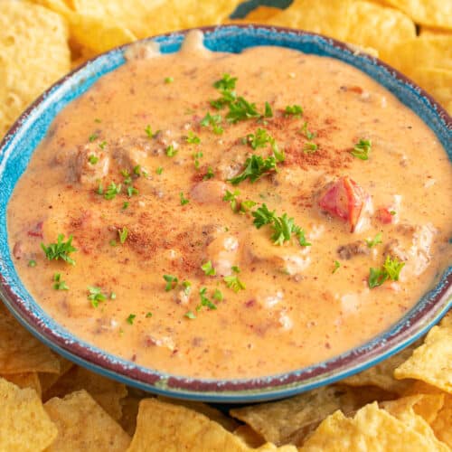 Bowl full of spicy sausage Rotel Queso Cheese Dip in a blue bowl surrounded by yellow tortilla chips.