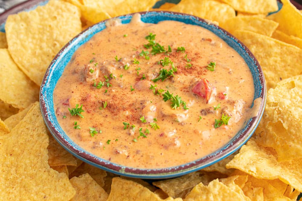 blue bowl full of spicy sausage rotel queso cheese dip on a plate of tortilla chips.