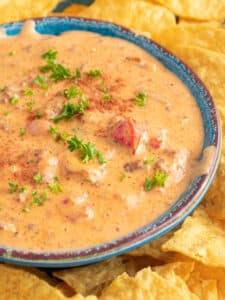 Bowl full of spicy sausage Rotel Queso Cheese Dip in a blue bowl surrounded by yellow tortilla chips.