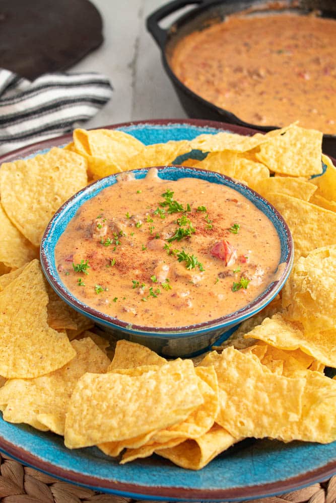 Blue bowl full of spicy sausage rotel queso cheese dip surrounded by tortilla chips in on a blue bowl with a skillet full of queso and black and white towel at a 45 degree angle.