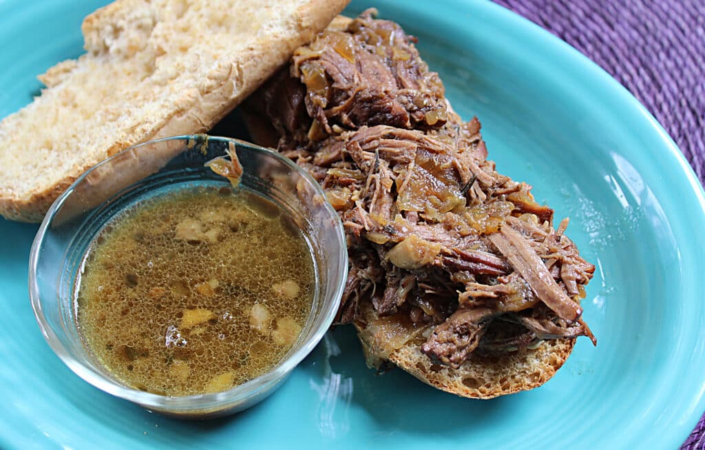 Drip beef sandwich open faced with a clear bowl of savory au jus on a blue plate.