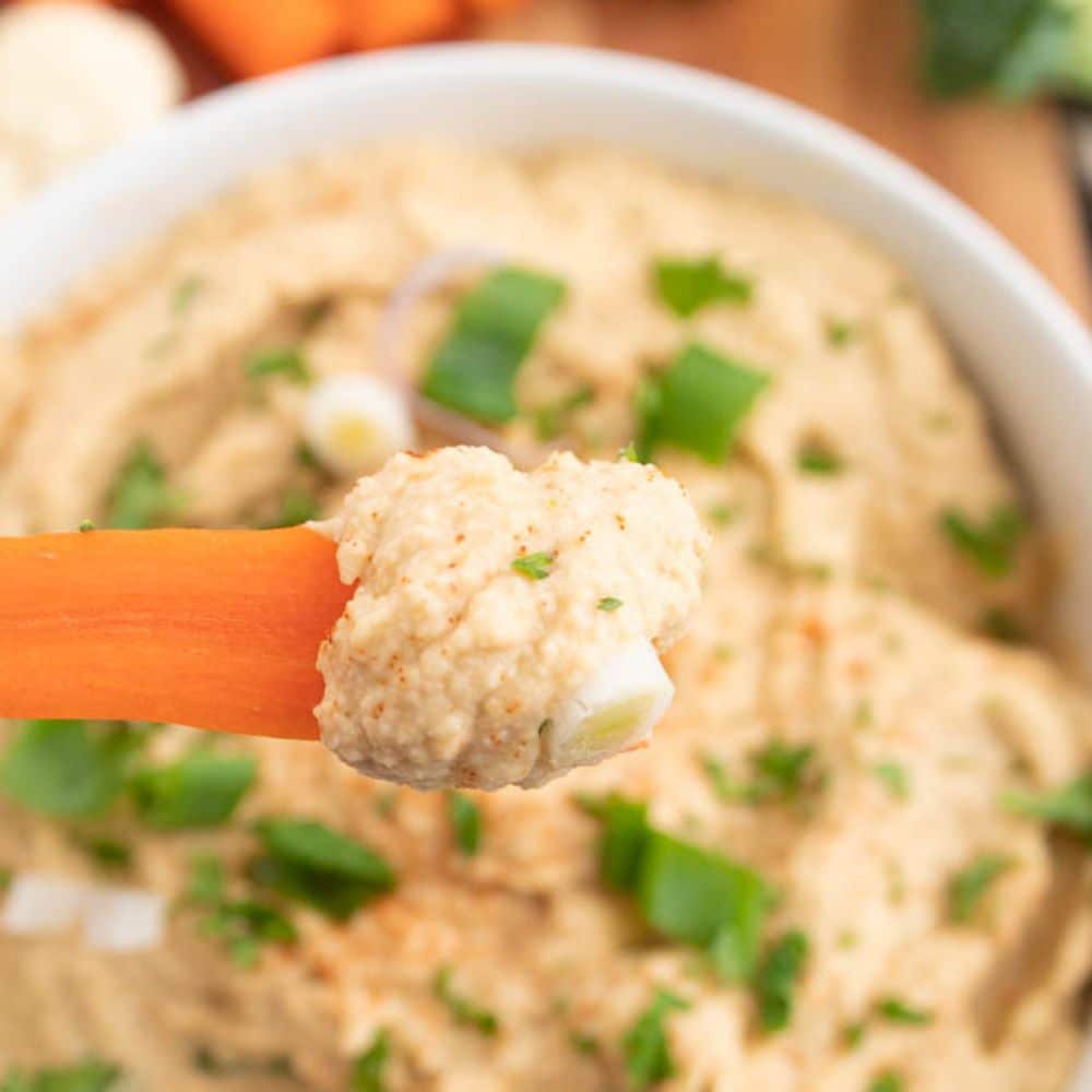 Basic homemade hummus serving on a carrot stick over a bowl of homemade hummus dip.