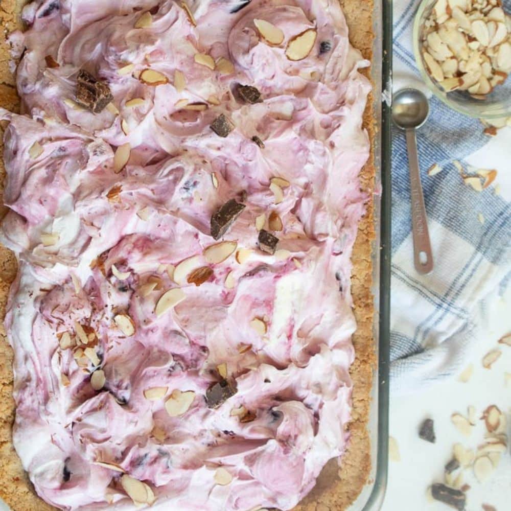 Easy creamy chocolate cherry fluff bars in a glass baking dish with sliced almonds and chocolate chunks on top. On a blue and white plaid towel.