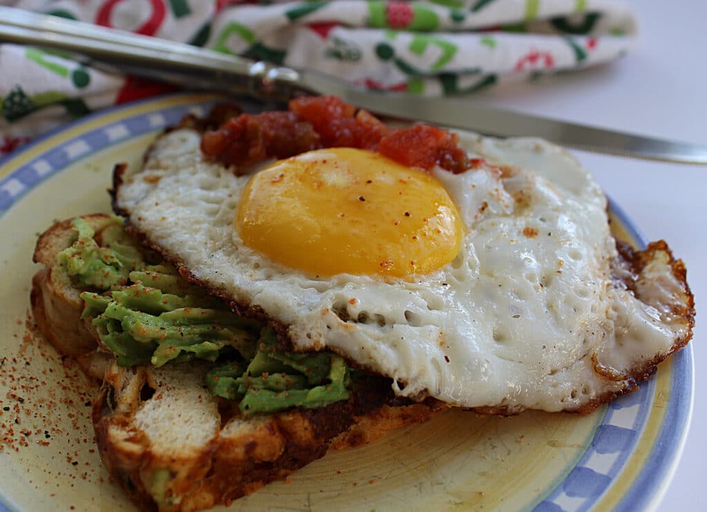 Fried egg on top of spicy avocado toast with a side of salsa on a blue and yellow plate with a knife on the side.