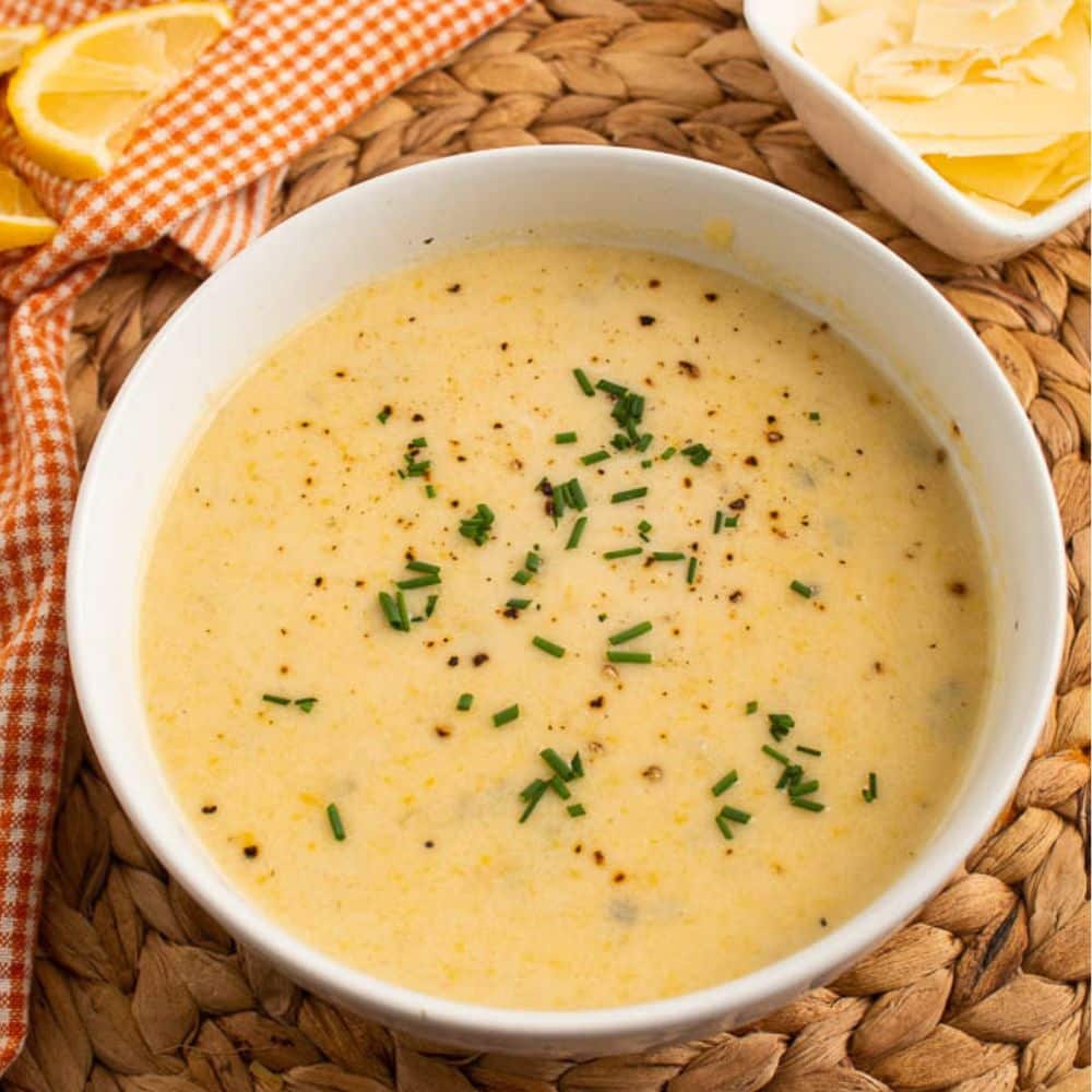 Creamy yellow summer squash soup with parmesan and lemon in a white bowl on a bamboo placemat and orange and white checkered towel with lemon wedge and extra parmesan.