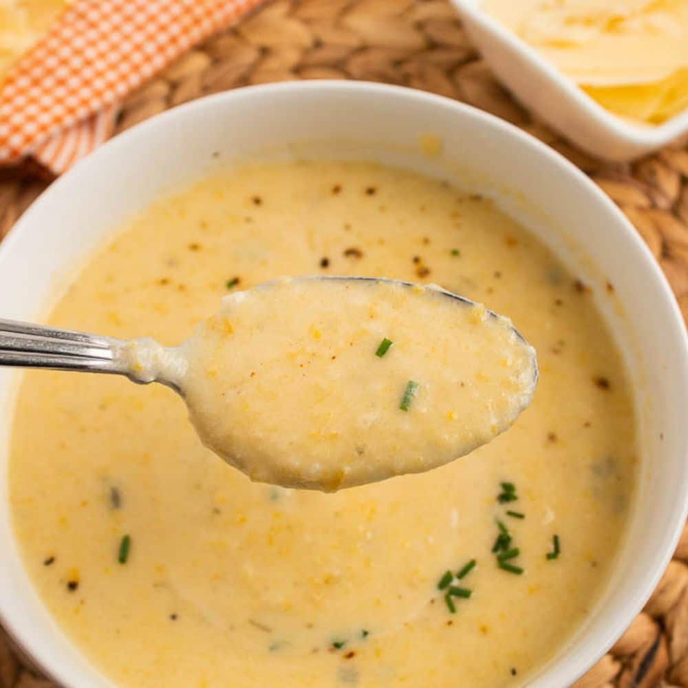 Spoonful of Creamy yellow summer squash soup with parmesan and lemon in a white bowl on a bamboo placemat and orange and white checkered towel with lemon wedge and extra parmesan.