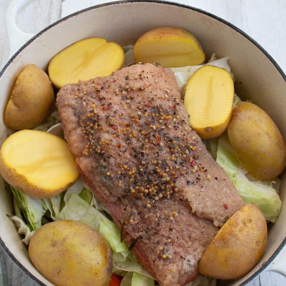 Boiled corned beef with seasonings and raw potatoes and cabbage in a white Dutch oven.