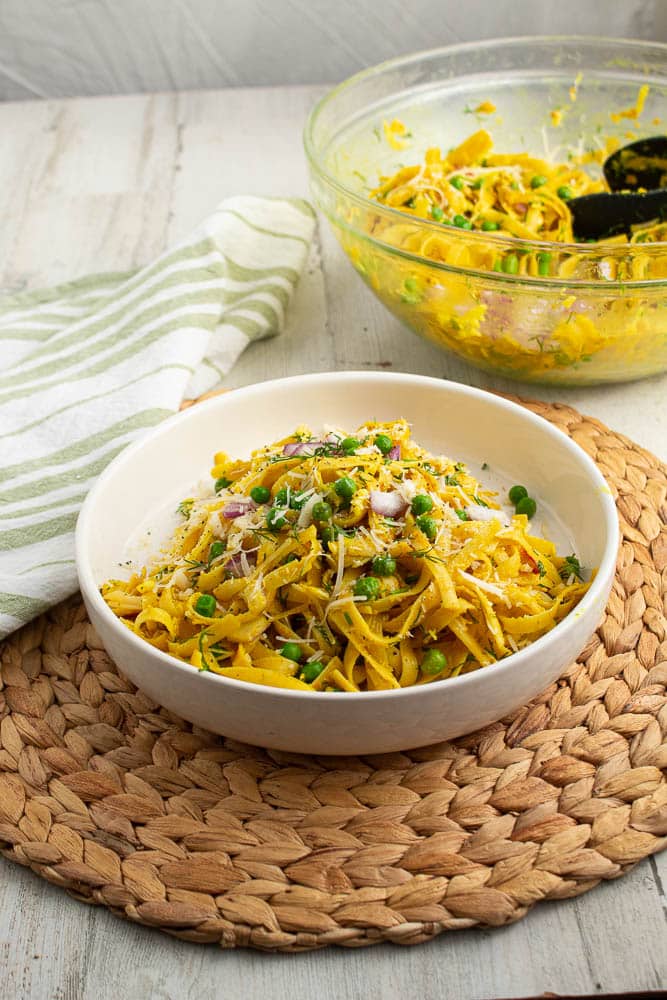 Curry pasta with peas, onions, fresh dill in a white bowl, on a bamboo placemat and a green and white towel. Large clear serving bowl in the background.